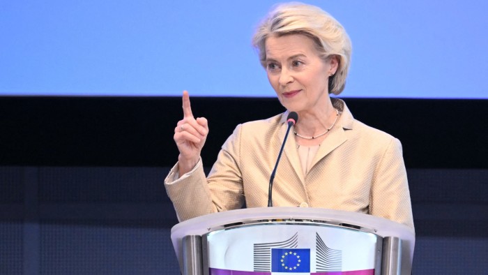 Ursula von der Leyen speaks at a podium with the European Commission emblem, raising one finger during a press conference.