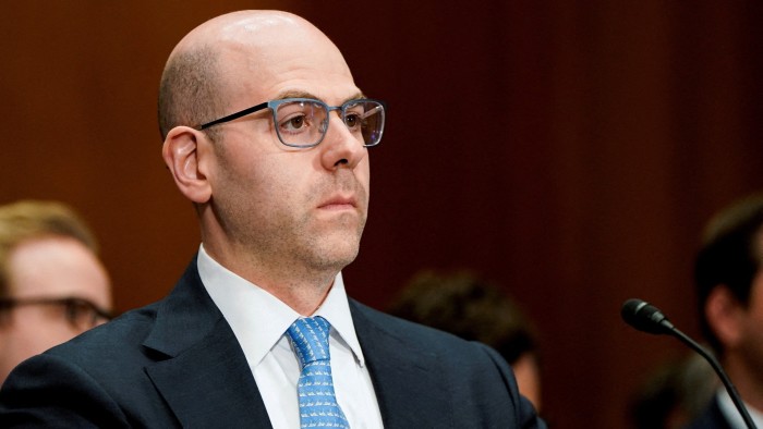 Stephen Miran appears seated and attentive during a Senate committee nomination hearing, wearing a suit and glasses.