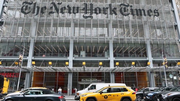 The front of The New York Times building with cars and a yellow taxi driving past on the street.