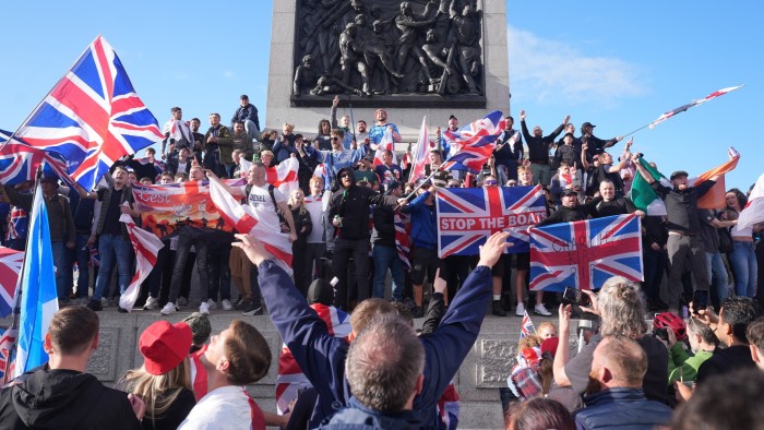 A large crowd at Trafalgar Square waving Union Jack flags and holding banners, including one reading "STOP THE BOATS," during a Unite the Kingdom rally led by Tommy Robinson.