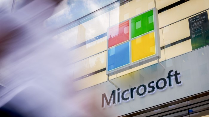 The Microsoft logo is displayed above the entrance of a Microsoft store, with motion blur from a passer-by in the foreground.