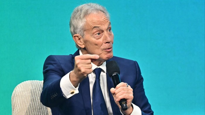 Tony Blair speaks into a microphone, gesturing with his hand during a panel discussion at the Austrian World Summit.
