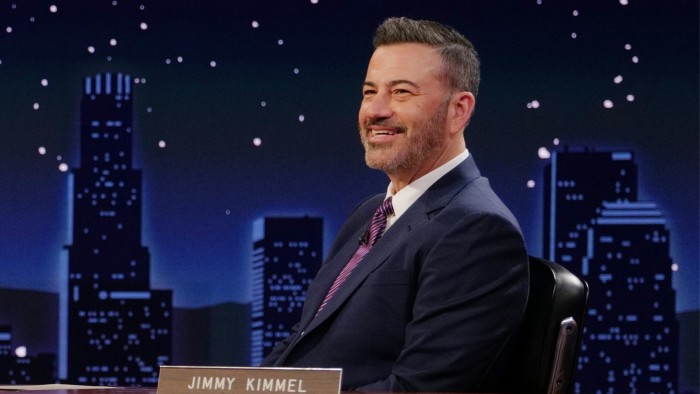 Jimmy Kimmel smiles while seated at his desk on the set of ‘Jimmy Kimmel Live!’ with a cityscape backdrop.