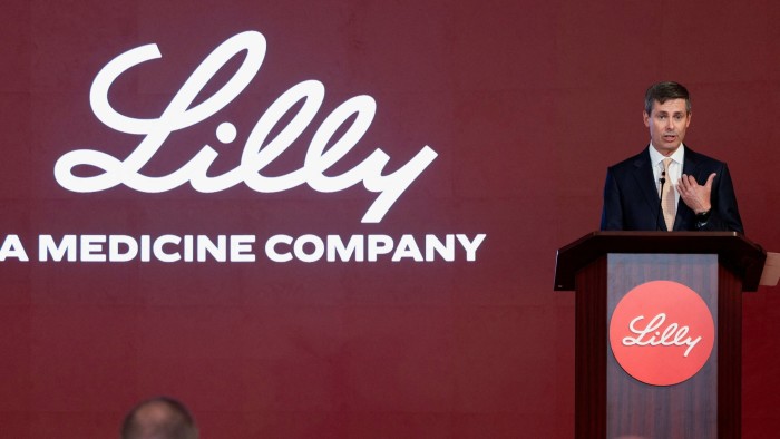 Dave Ricks speaks at a podium during an Eli Lilly press conference, with a large Lilly logo displayed behind him.