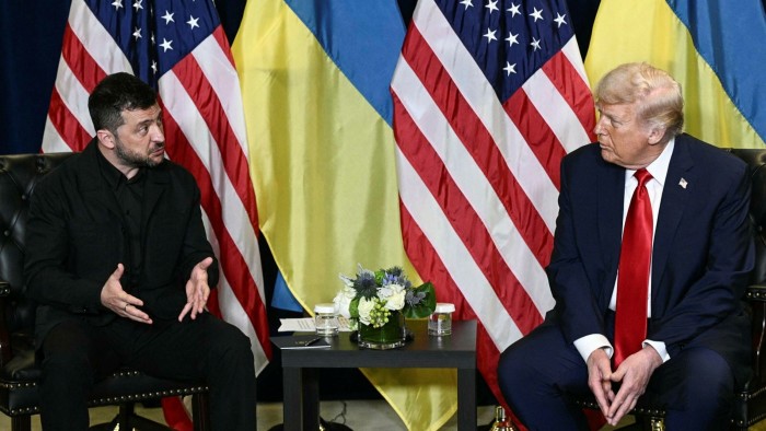 Volodymyr Zelensky gestures while speaking to Donald Trump during a meeting, with US and Ukrainian flags behind them.