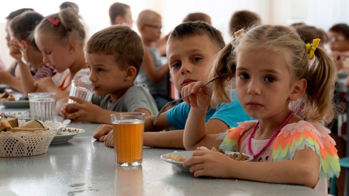 Children sit closely together at a table eating a meal, with a girl in colorful clothing looking toward the camera.