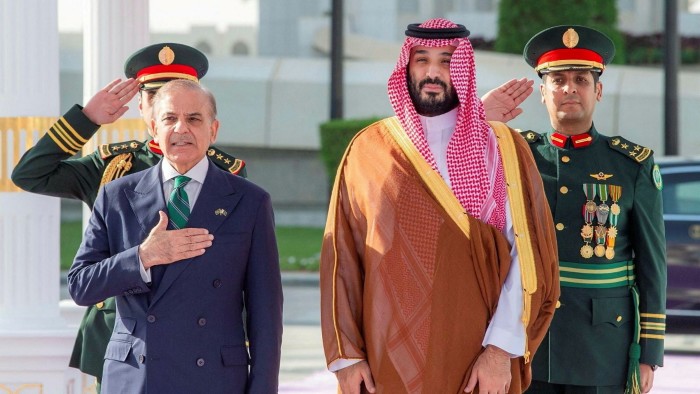 Shehbaz Sharif stands with his hand on his chest next to Mohammed bin Salman, who wears a traditional Saudi robe, as two uniformed officers salute behind them.