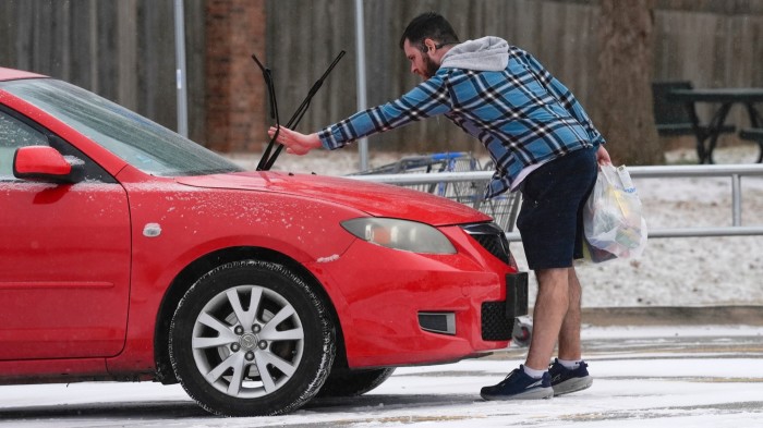 A man lowers the windshield wipers on his red car while holding groceries in a snowy parking lot during a winter storm.