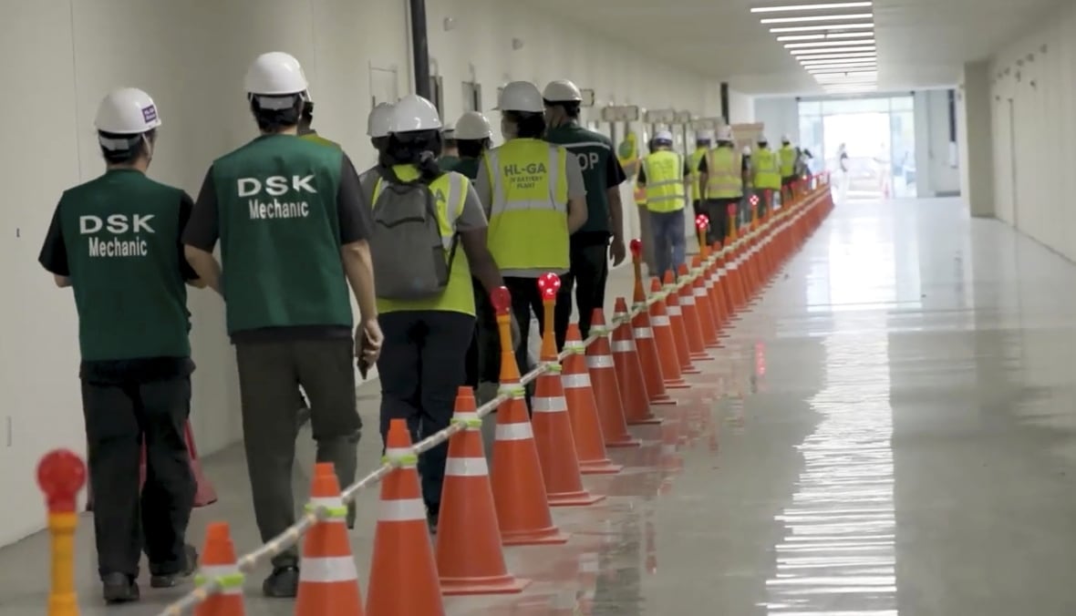 A group of people walk down a hallway single file, with a line of orange pilons beside them. They are all wearing hard hats and most are in yellow vests, with two workers at the back of the line wearing green vests that say "DSK mechanic" on them. 