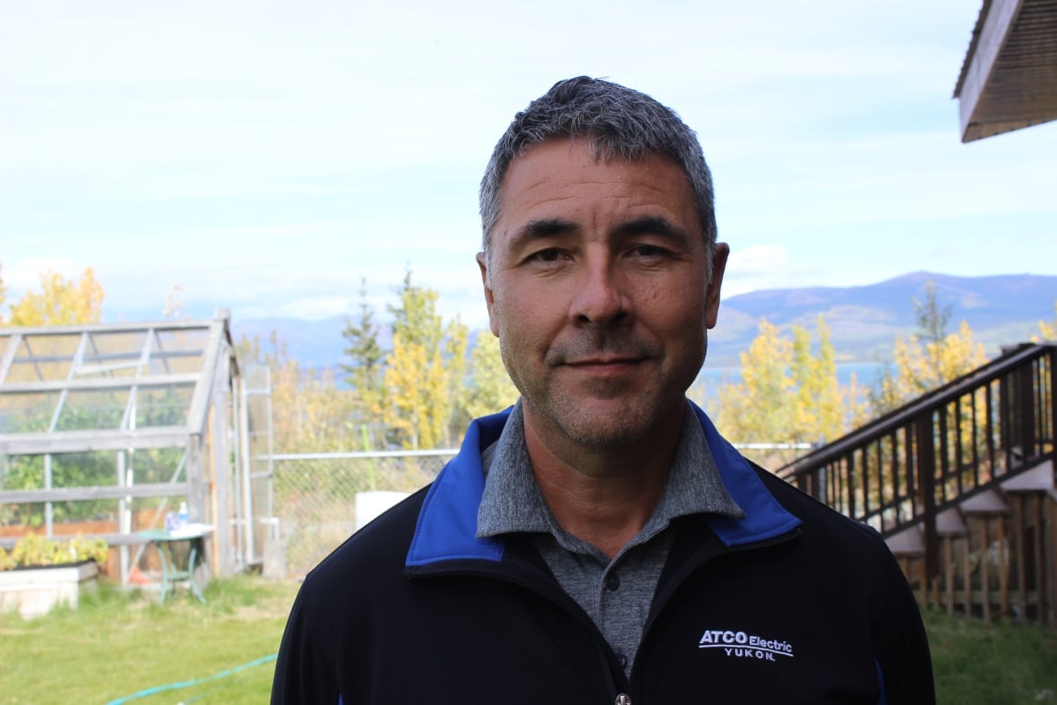 A man in a black jacket stands in a yard before a backdrop of mountains.