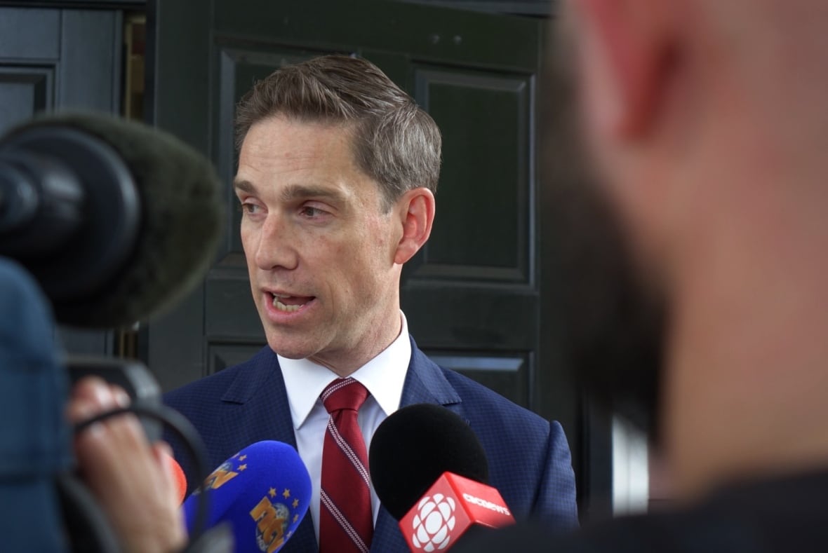 A man wearing a suit stands in front of a large black door speaking with reporters.