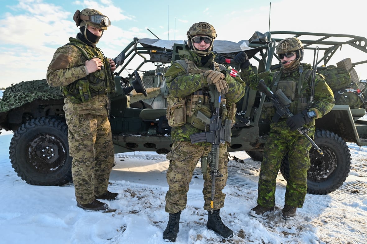 Three men in green army uniforms stand in front of a military vehicle.