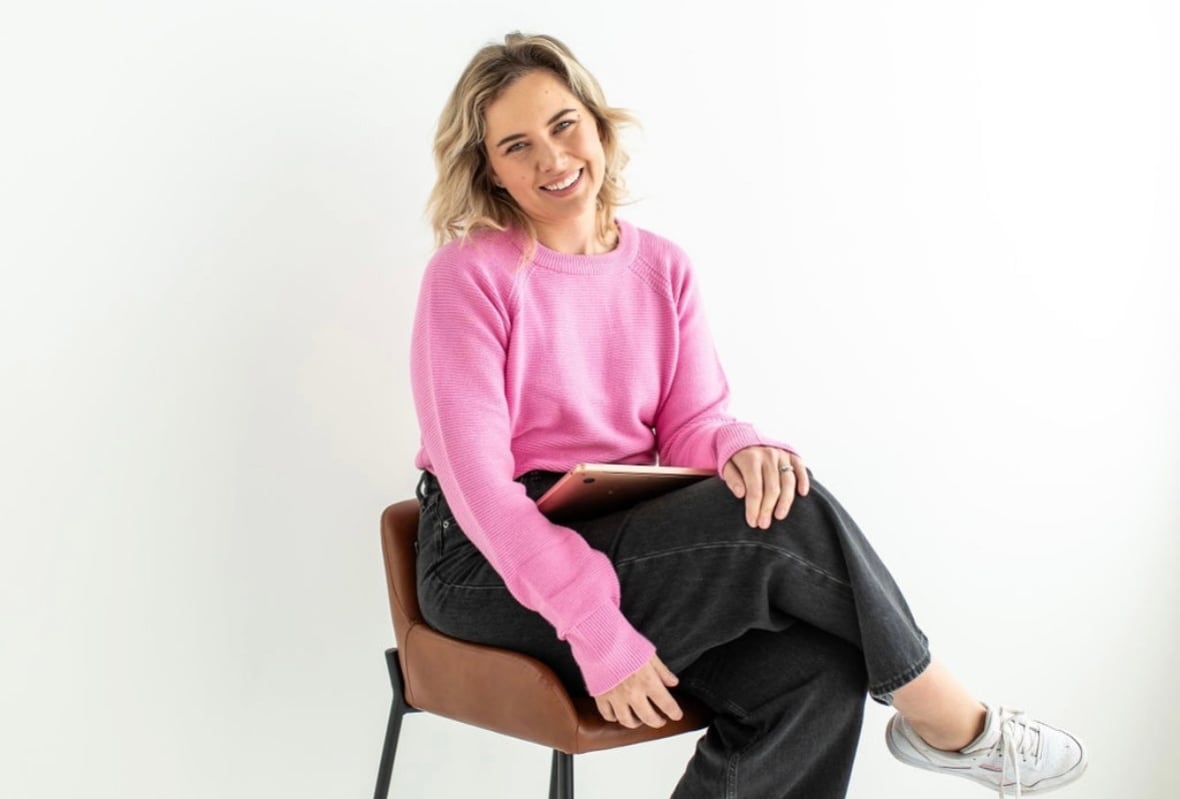 A woman in a sweatshirt poses for a photo on a stool in front of a white background