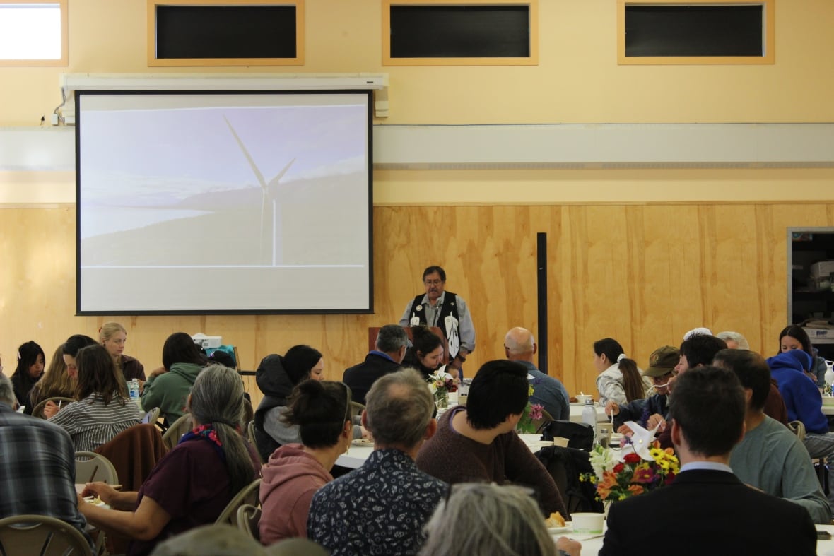 A wide shot with a bunch of people at tables in the foreground, and a man standing at a podium in front of them.