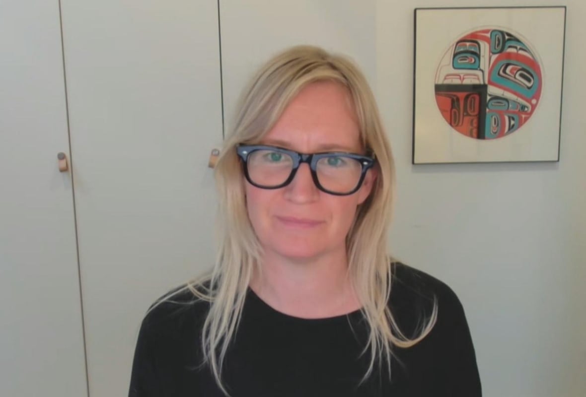 A white woman smiles in front of a closet and a painting with Coast Salish art.