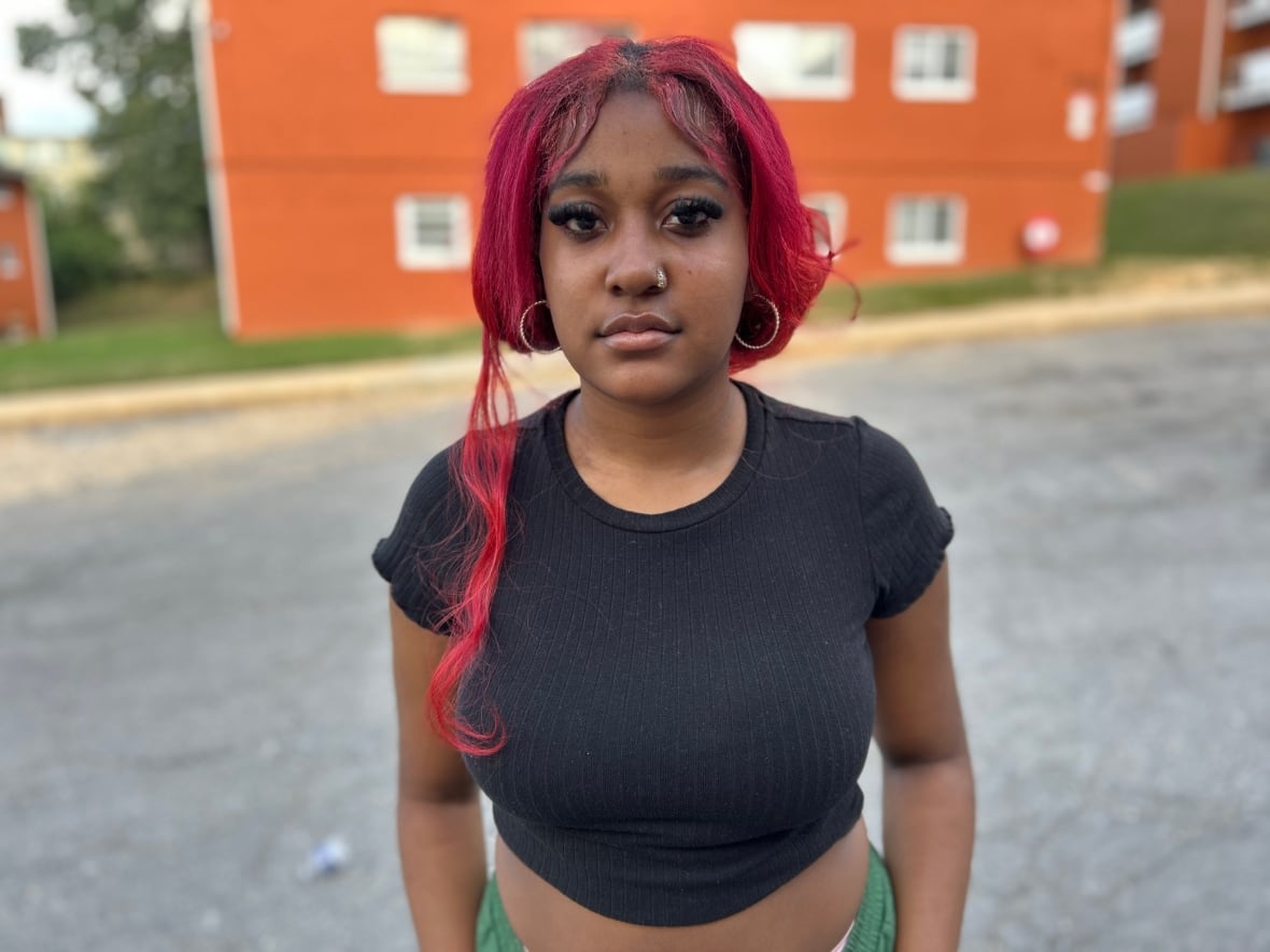 Laniyah King is 17. She has fuchsia coloured hair tied back in a low ponytail. Her bangs are parted in the middle. She's wearing a black crop top and green pants. She is standing in front of a red brick building.