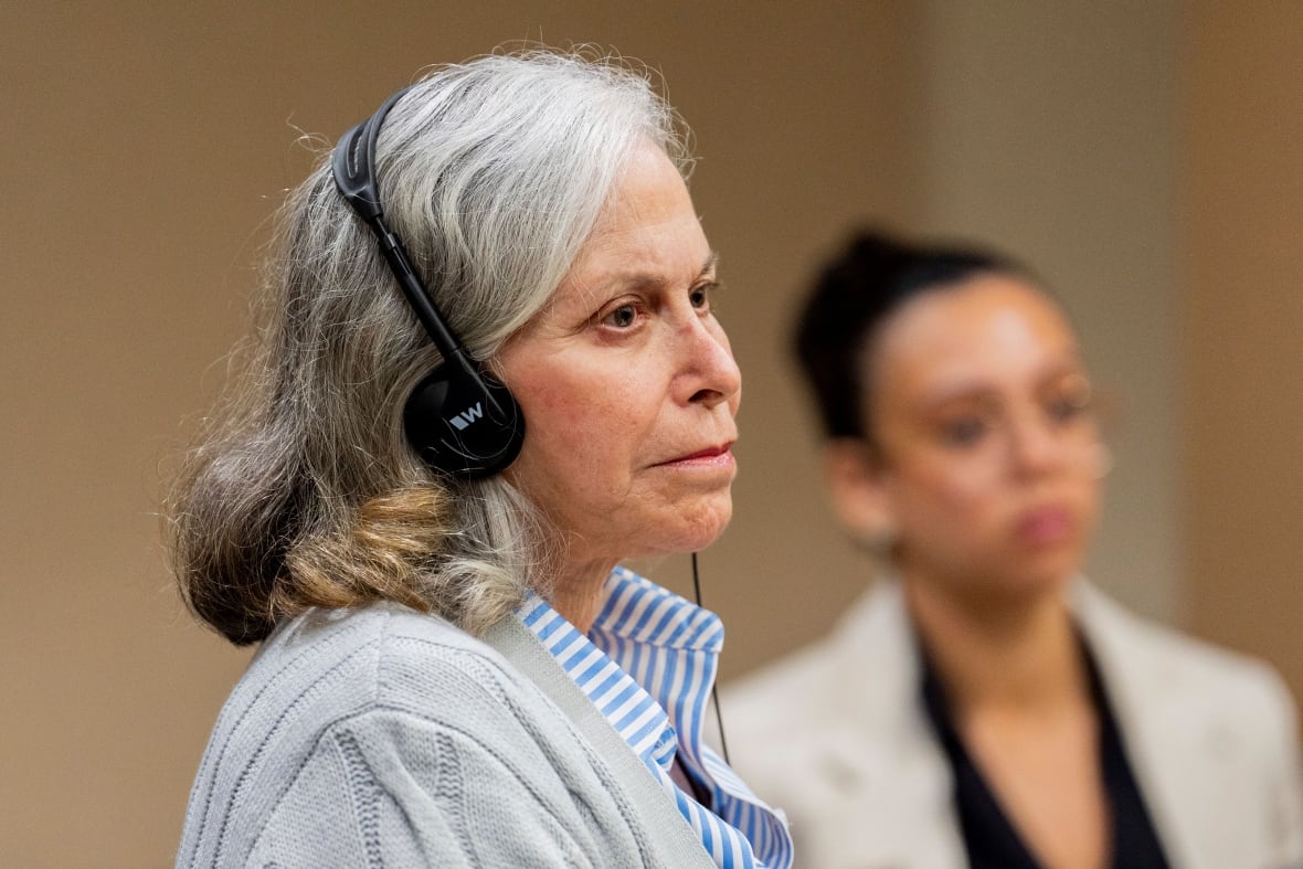 Grey haried woman in a shirt and cardigan sits and listens in court, wearing headphones