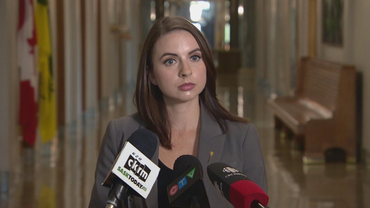 a woman in a grey blazer and black shirt stands in front of three microphones. 