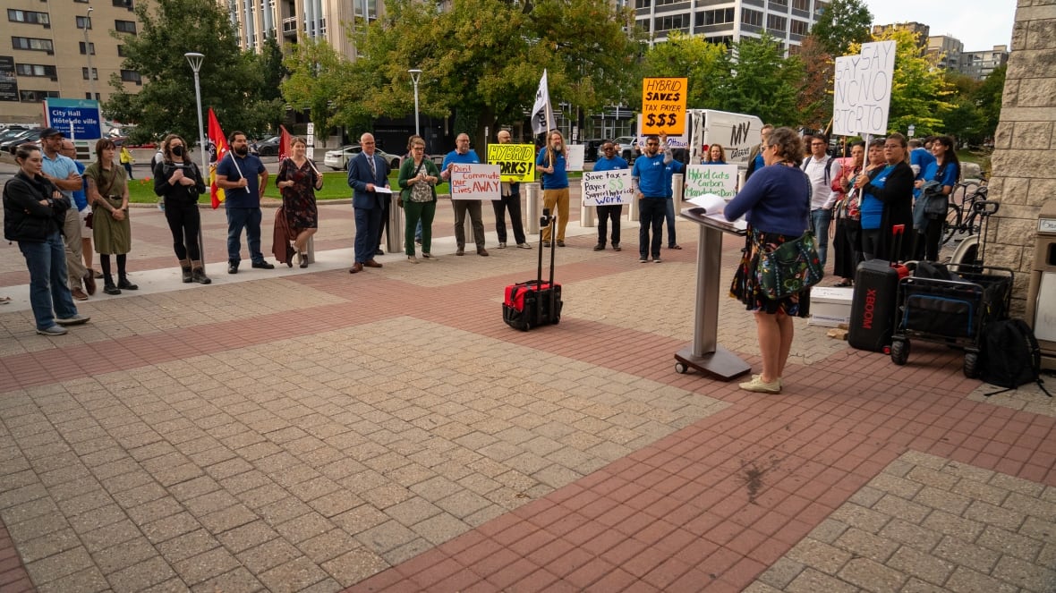A small crowd of people hold signs and listen to a speaker at an outdoor rally in autumn outside a city hall.