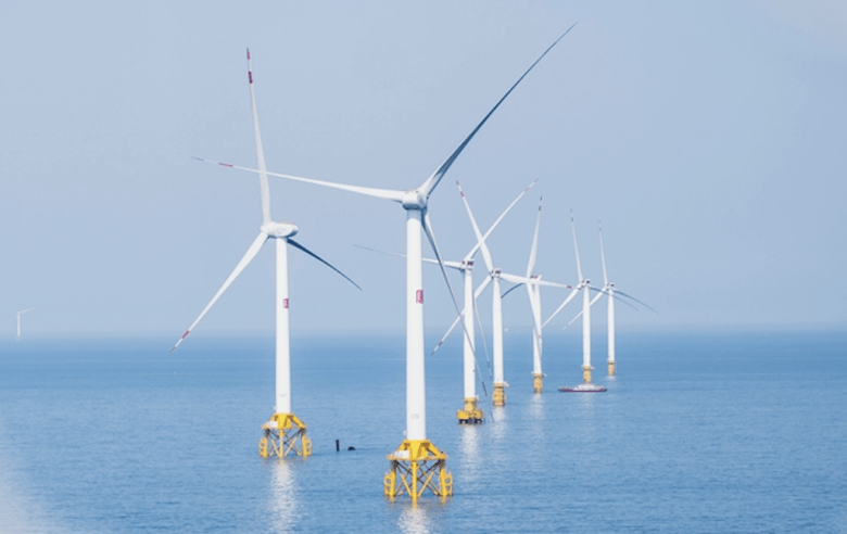 A row of white wind turbines perched on bright yellow bases in the middle of the ocean on a calm day.