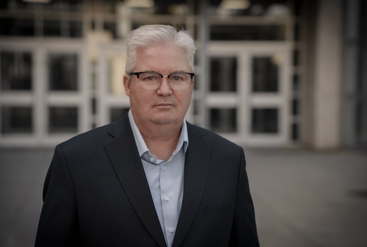 A man with short, white hair and black, rectangular glasses wearing a blue collared shirt and dark blazer stands in front of glass doors looking at the camera. 