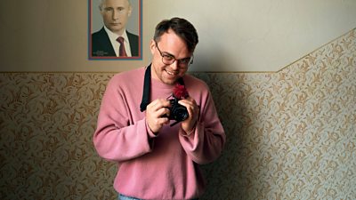 A man in a pink sweatshirt holds a camera in front of some patterned wallpaper.  There is a photo of Putin in a frame also on the wall.