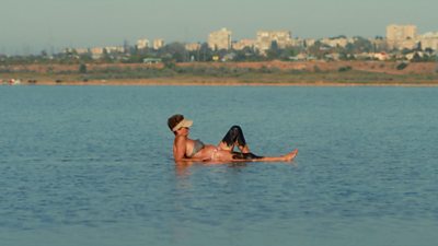 A woman wearing a bikini and a visor lying in a shore of shallow water. She has black mud on her legs.