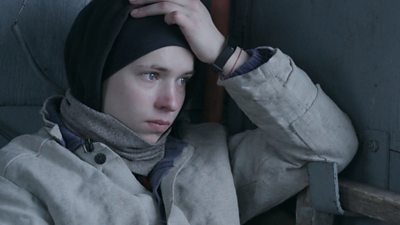 A young woman wrapped up in a grey winter coat and black headscarf resting her head on her left hand.  She looks upset.