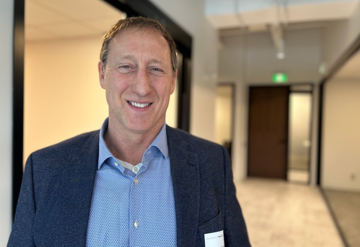 A man with short brown hair smiles at the camera, while wearing a blue shirt and suit jacket, standing in a hallway.