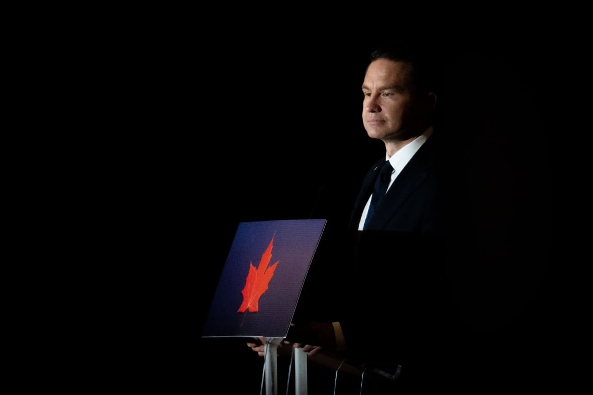 A man in a dark suit stands behind a podium with a blue placard featuring a red leaf.