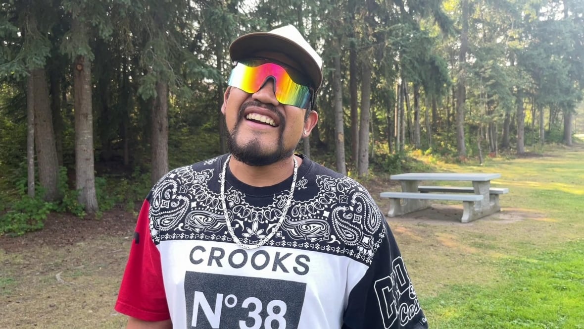 A man laughs while wearing aviators and a ballcap in a park.