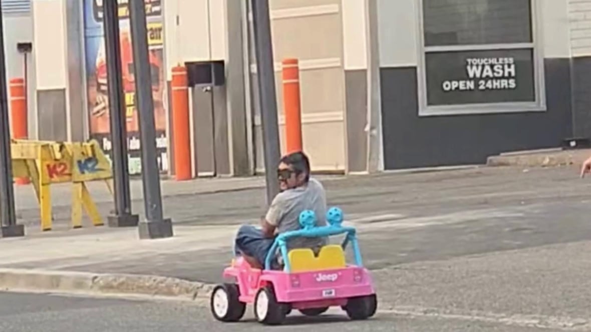 A man drives a child-sized pink Barbie Jeep along a road.