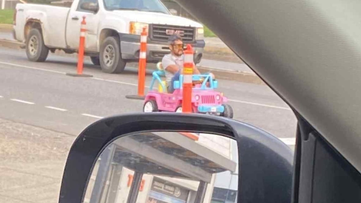 A man driving a toy pink Barbie Jeep as viewed from a vehicle window.