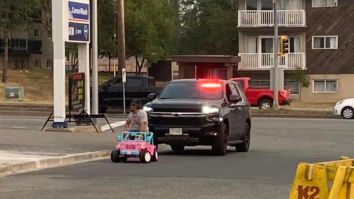 A man in a child-size pink Barbie Jeep is pulled over by an unmarked police cruiser with its lights on.