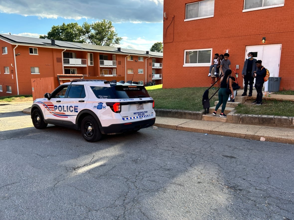 The photo shows a Metropolitan Police Department SUV driving by a group of teens standing on the steps of an apartment building in Washington. There are two red brick buildings in the photo. 