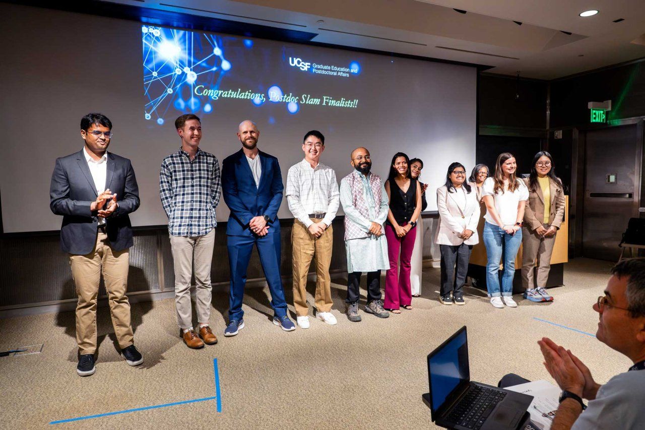 Presenters line up on stage in front of a projector screen that reads "Congratulations, Postdoc Slam Finalists!"