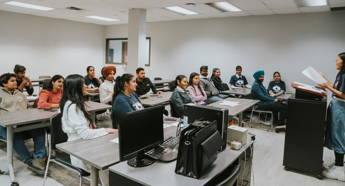A class of sitting adult students look at a standing teacher.