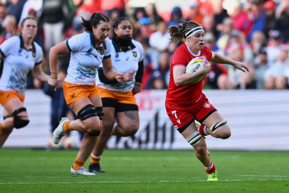 A female rugby player representing Canada runs with the ball in her right hand as Australian opponents trail behind during a day match.