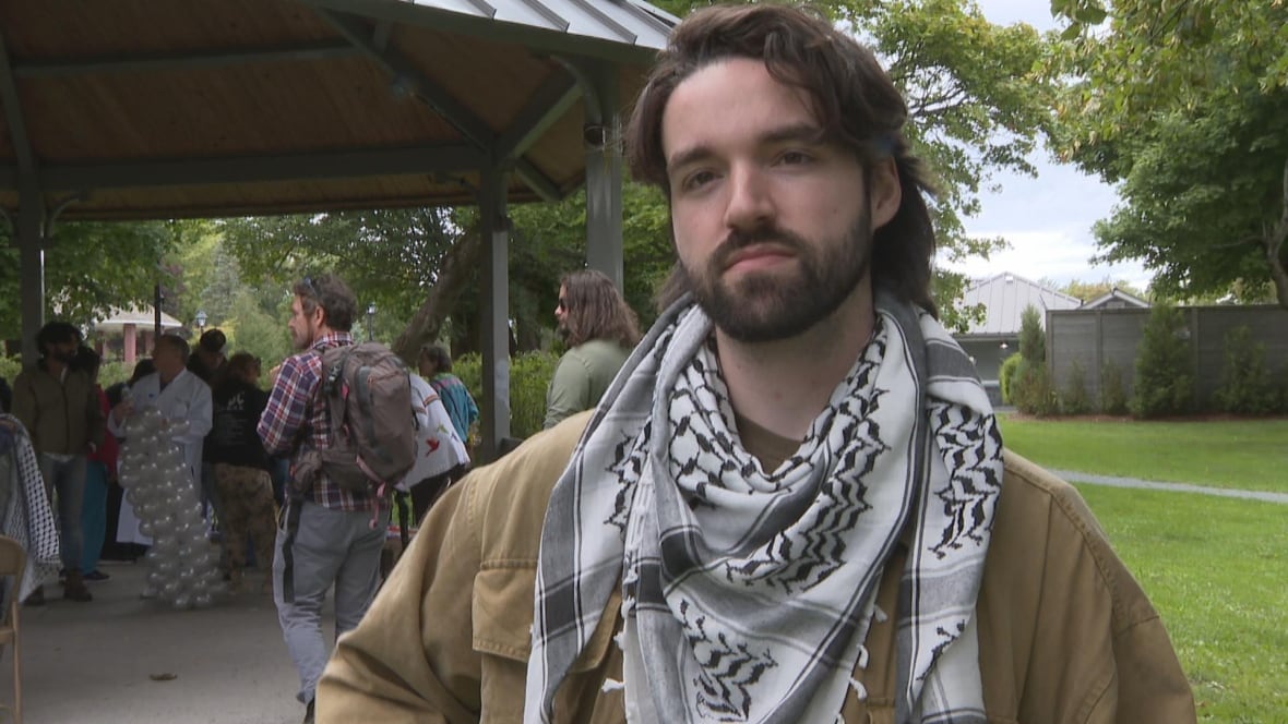 A brunette with a keffiyeh scarf and yellow jacket stands in a park, looking toward the camera.
