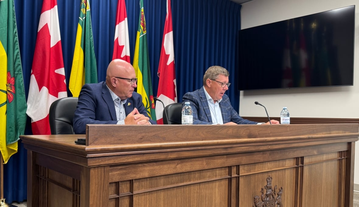 Two men in suits sit at a dark brown large desk, with Saskatchewan and Canada flags behind them.