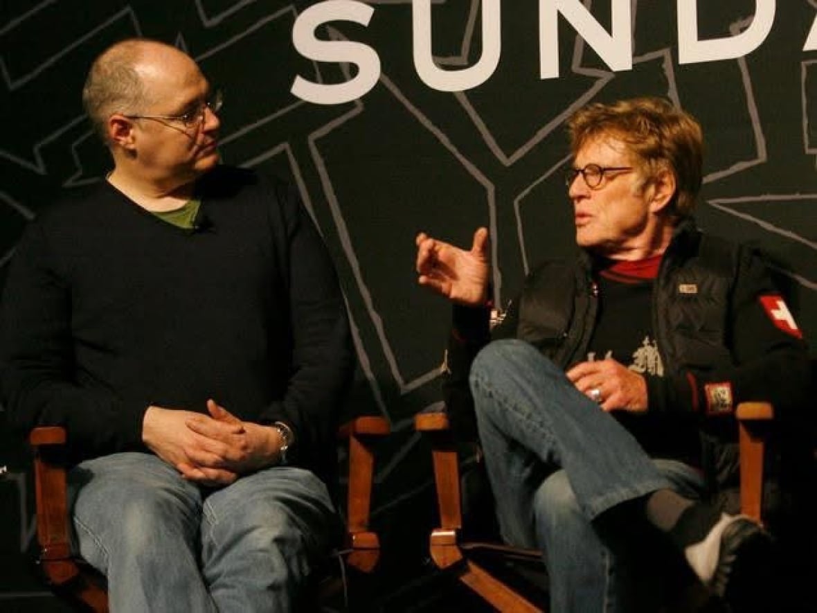 two men sit next to eachother in fold out directors chairs, speaking to one another. both are wearing jeans and seem to be engaged deeply in the conversation.