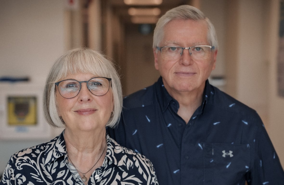 a woman in a black and white shirt with grey hair stands on the left, and a man with a blue shirts stands behind her to her right