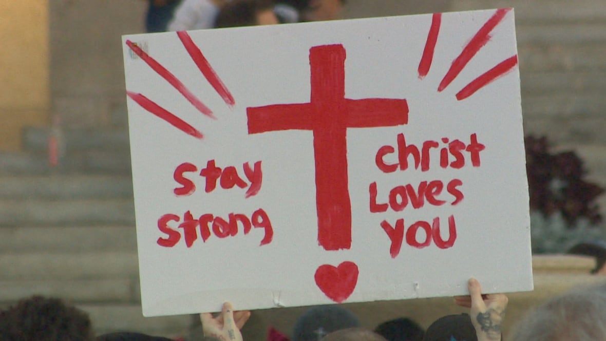 a sign is held up in a crowd of people gathered outside. It says "Stay strong, Christ loves you."