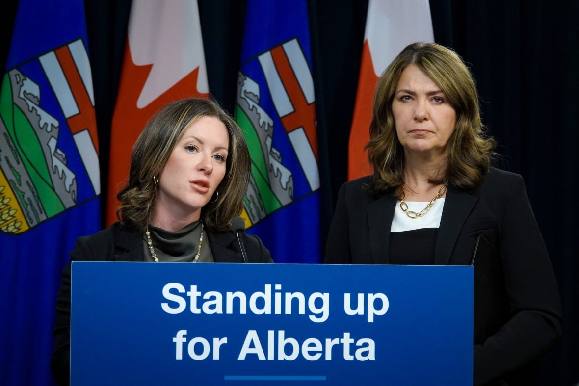Two woman stand at a podium, with a sign stating Standing Up for Alberta