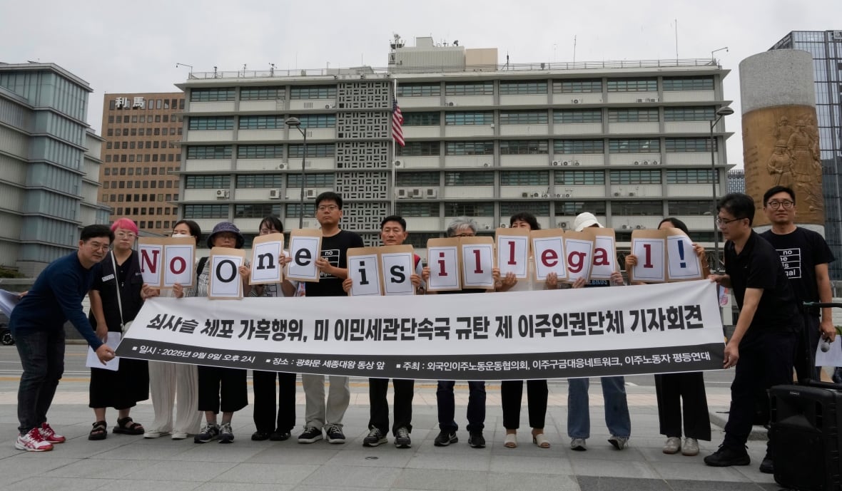 A group of people hold a sign up with Korean letters on it while others hold single letters above that spell out "No One is Illegal" in English.  