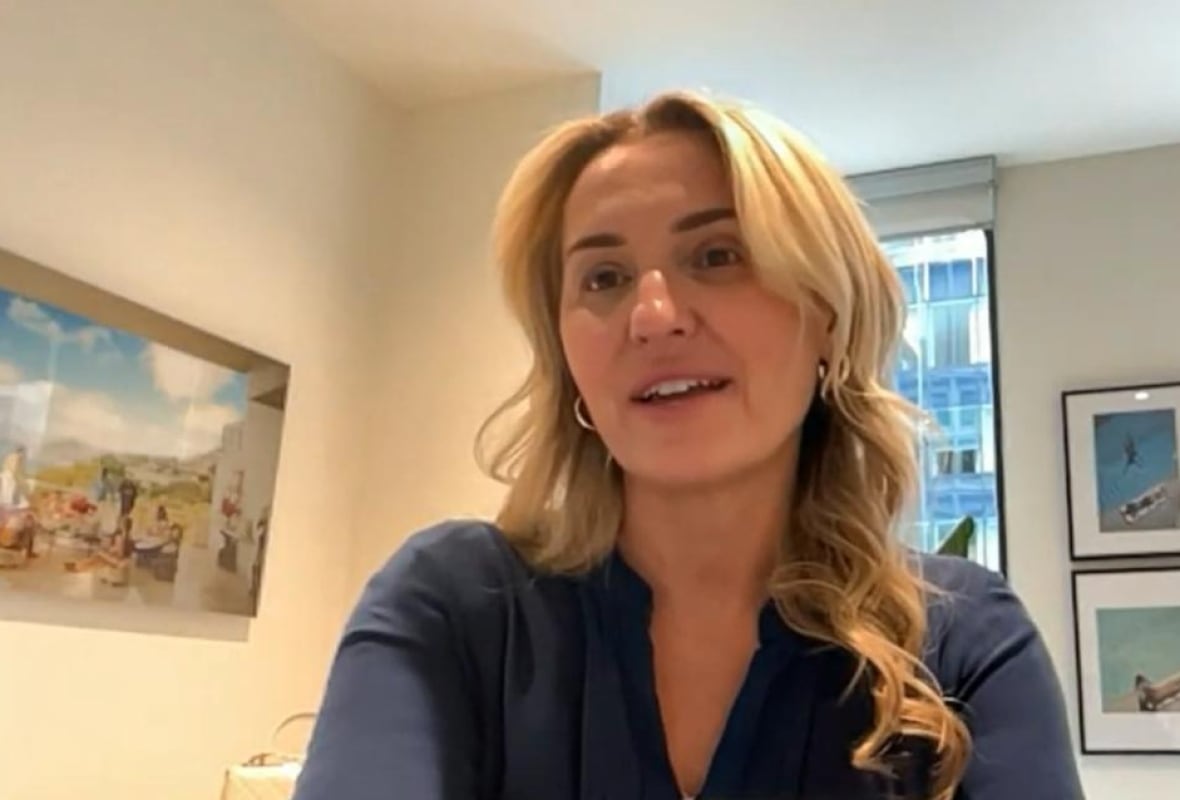 A blonde woman sits in an office with pictures on the wall and a window behind her.