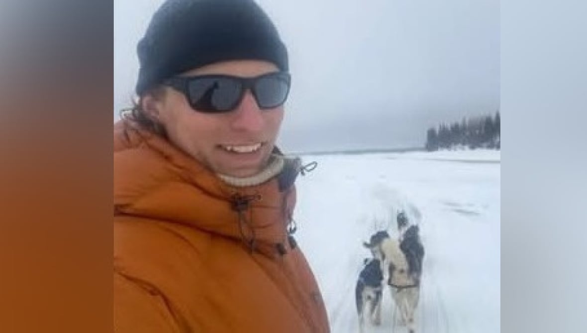 A man wearing a black tuque and orange parka and black sunglasses looks towards the camera. It's snowy outside. There are dogs in the background.