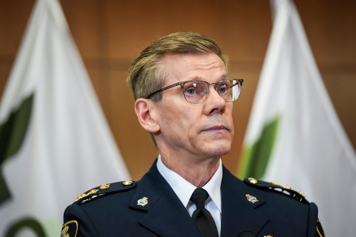 A man in a police uniform, wearing glasses looks forward with a stern expression on his face.