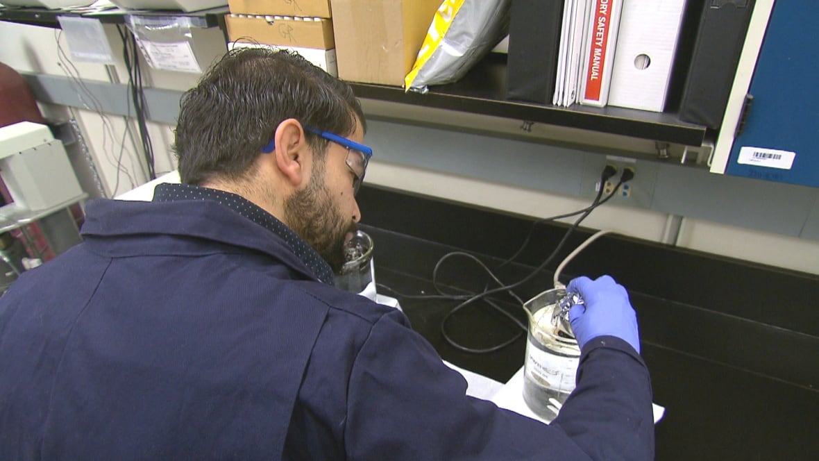 A man wearing a blue lab coat and goggles leans over a desk, with his gloved hand hovering over a beaker filled with liquid. 
