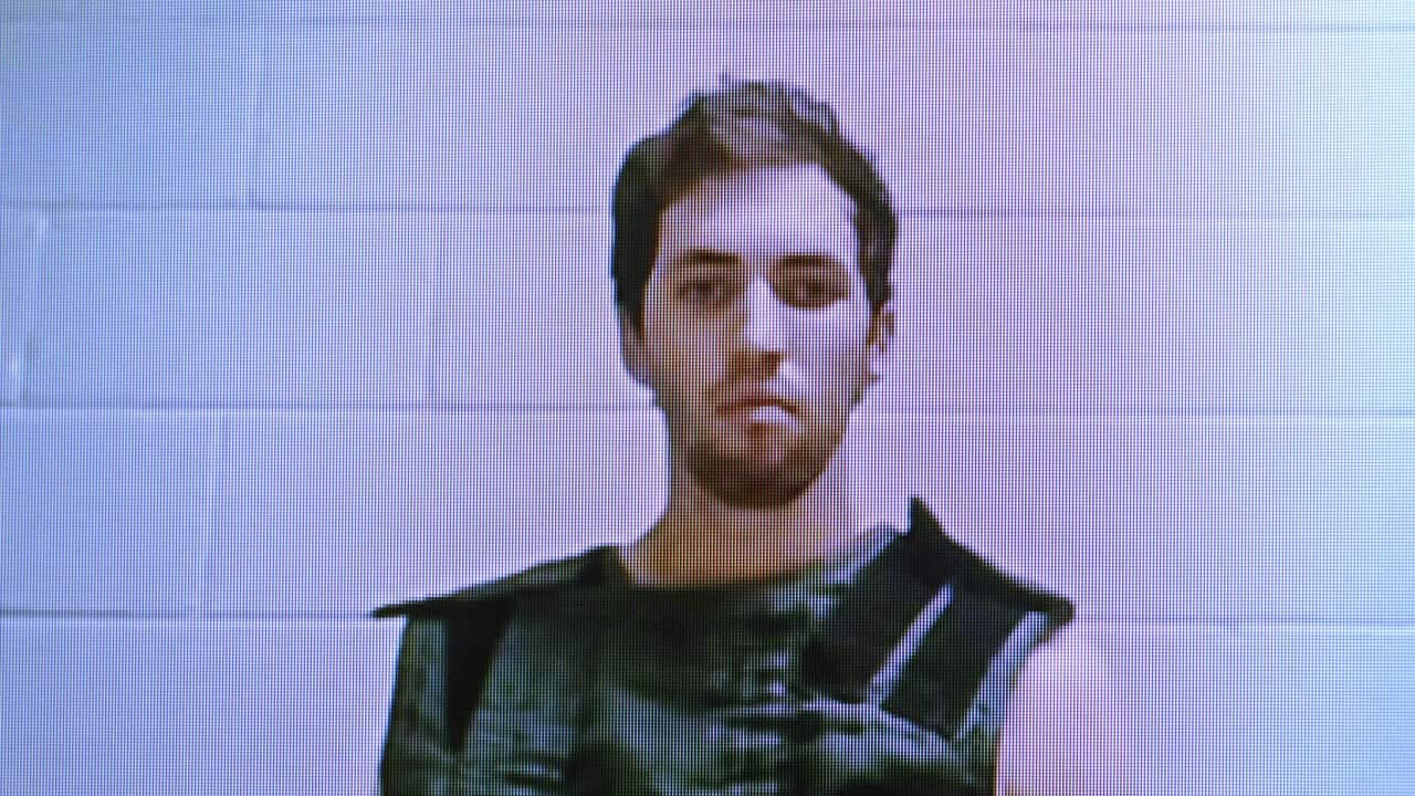 A young man with short brown hair and a beard wearing a prison vest seated in front of a white brick wall.